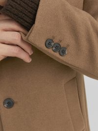 Smeđi vuneni kaput s krojenim fitom, s izrezanim ovratnikom i tri crna gumba na manžetama uz pletenu detalj s rebrastim uzorkom ispod.