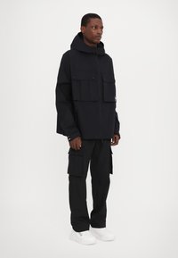 Black hooded jacket with large flap pockets, paired with black cargo pants and white sneakers. Features a zip closure and minimal branding.