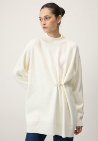 Valge oversize kampsun poolkaelusega, millel on kogutud detail, mis on kinnitatud metallkinnitusega. Pehme tekstuur, pikad varrukad ja soonikuga ääred.