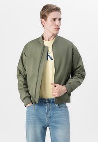 Oliven grønn bomberjakke med glidelås, ribbestrikkede mansjetter og sidelommer, iført over en gul T-skjorte og blå denimjeans.