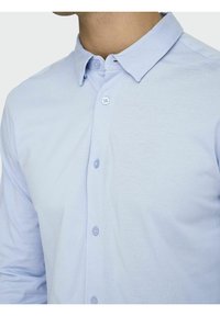 Chemise bleu clair à boutons, en tissu doux ; présente un col pointu et deux boutons sur la patte de boutonnage avant. Texture lisse.