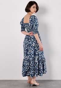 Robe maxi floral bleu et blanc, avec un décolleté carré, des manches bouffantes, une taille élastique et un ourlet à volants. Texture de tissu lisse.