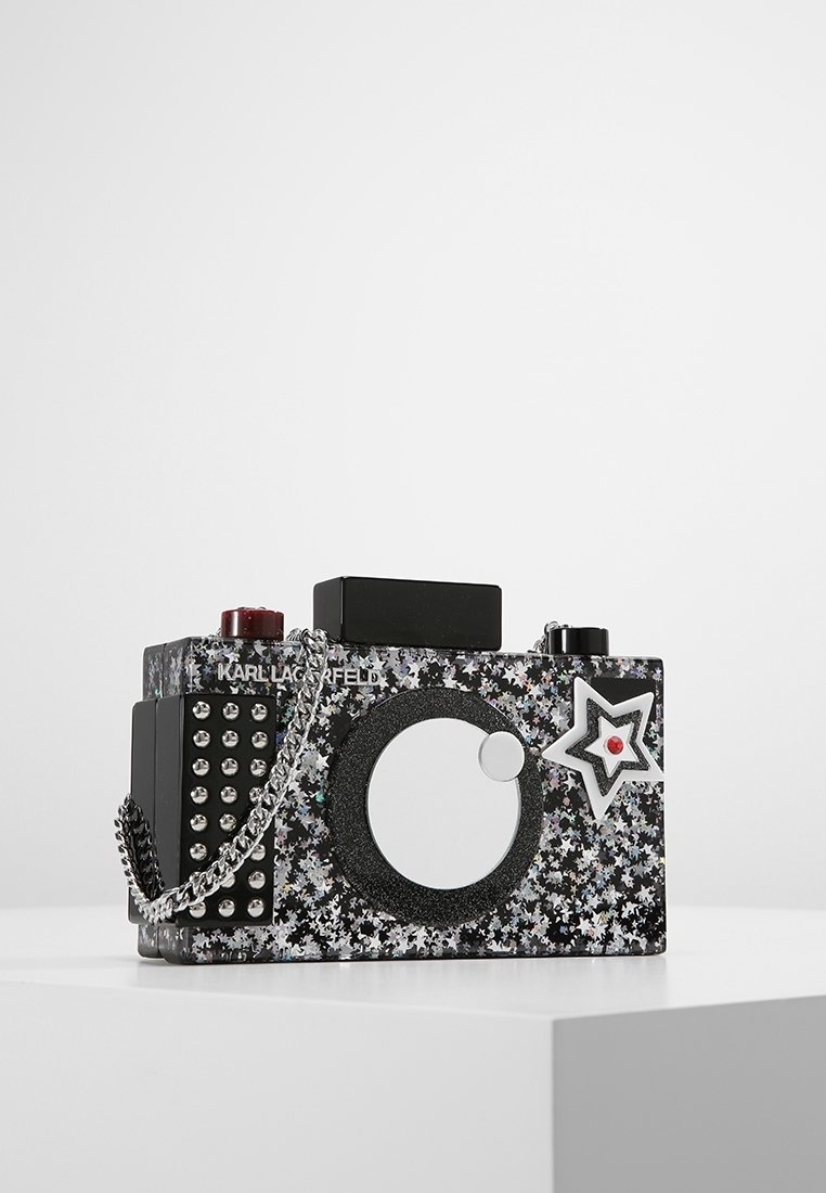 Pochette en forme d'appareil photo noire et argentée à motif étoiles, avec bandoulière chaîne, détails cloutés et décoration étoile sur le devant.