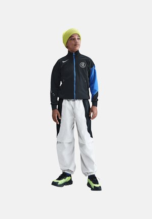 Giovane in piedi che indossa una giacca con cerniera nera e blu, pantaloni della tuta bianchi e neri, sneakers verde neon e nere e un berretto lavorato a maglia giallo brillante.