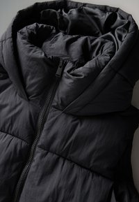 Fekete pufi kabát, magas gallérral és zipzáras lezárással. Az anyag texturált, varrott szakaszokkal a szigetelés érdekében.