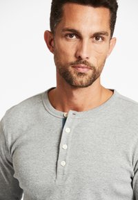 Graues Langarm-Henley-Shirt mit rundem Ausschnitt, fünf cremefarbenen Knöpfen und einer dezenten Textur; kurze Nähte am Kragen.