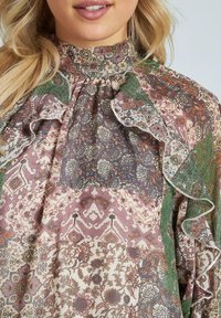 Blusa multicolore con motivi floreali e geometrici, colletto alto a volant e tessuto trasparente con tonalità di verde, rosa e beige.