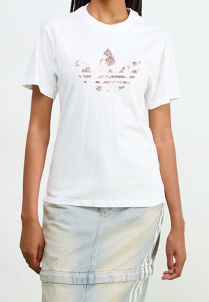 Osoba nosící bílé tričko Adidas s leopardím vzorem loga a světle modrou džínovou sukní s bočními pruhy.