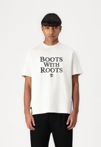 Krém színű, rövid ujjú póló, amelyen nagy, fekete felirat olvasható: "BOOTS WITH ROOTS", valamint egy kis logó alatta. Fekete nadrággal viselve.