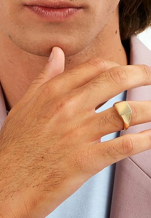 Gold-toned metal ring with a geometric, triangular shape and a polished surface, worn on a hand with light hair and a muted pink jacket.