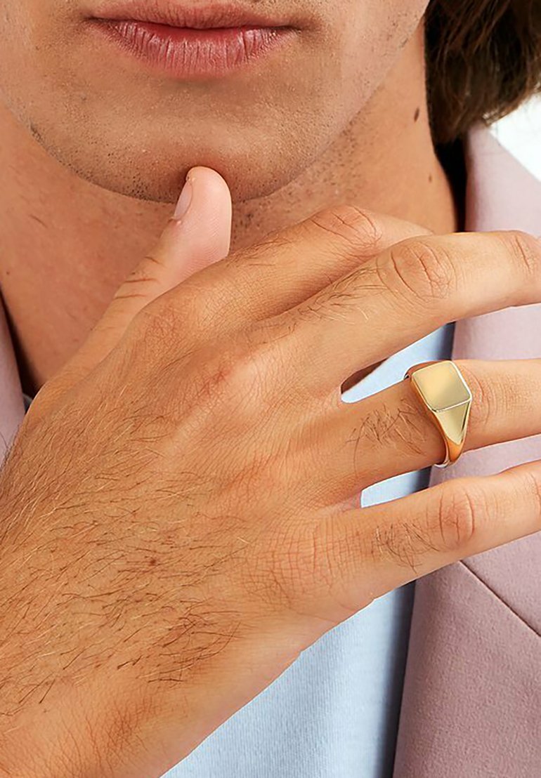 Goldfarbener Metalling mit einer geometrischen, dreieckigen Form und einer polierten Oberfläche, getragen an einer Hand mit hellen Haaren und einer gedämpften rosa Jacke.