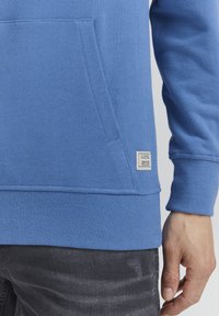 Sudadera azul de manga larga, con puños y bajo acanalados, bolsillo tipo canguro, textura suave, que presenta una etiqueta en el lado inferior izquierdo.