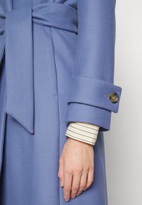 Abrigo azul claro con un acabado texturizado, con cintura atada, puños anchos y un detalle de botón. Debajo, una camisa blanca con rayas azules.