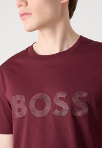 Bordová bavlnená tričko s potlačou loga "BOSS" v tmavšom odtieni na mriežkovanom vzore, s okrúhlym výstrihom a krátkymi rukávmi. Hladká textúra.