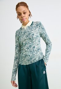 Osoba u dugom rukavu sa cvjetnim uzorkom, bijelom kragnom i tamnozelenim Adidas hlačama, stoji ispred jednostavne pozadine.