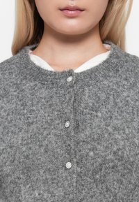 Grijze gebreide cardigan met een ronde hals, voorzien van drie glanzende knoopsluitingen en een zachte, textuurstof. Onder de cardigan is een wit kledingstuk zichtbaar.