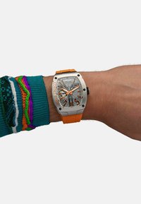 Armbanduhr mit orangenem Silikonarmband, silbernem Metallgehäuse, grauem Zifferblatt, schwarzen Akzenten und auffälligen orangen Stundenmarkierungen. Bunte gestrickte Ärmelsichtbar.