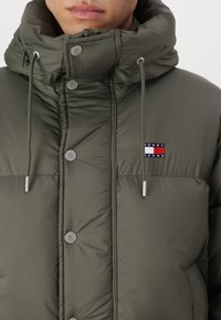Chaqueta acolchada verde con un alto cuello, cordón, botones plateados y un pequeño parche de logo en azul, rojo y blanco en el pecho.
