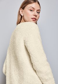 Sweter w kolorze kremowym, o puszystej teksturze, z okrągłym dekoltem i opuszczonymi ramionami, noszony przez modelkę, która spogląda przez ramię.