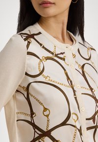 Cardigan cu nasturi, cu un fundal crem, decorat cu un model de lanț în tonuri de maron și auriu. Material texturat cu un finisaj moale.
