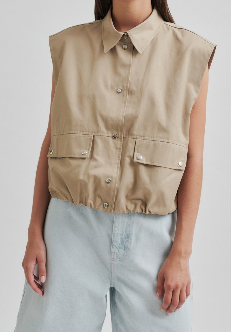 Veste beige sans manches à boutons avec poches avant portée sur un jean bleu clair taille haute sur une personne aux cheveux mi-longs.