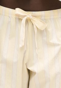 Pantalon de pyjama beige clair avec de fines rayures verticales et un nœud en tissu noué à la taille élastique.