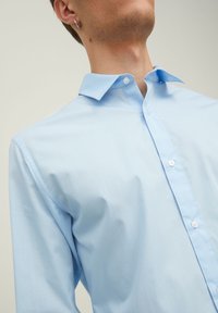 Light blue button-up shirt with a classic collar, smooth texture, and white buttons, fitted at shoulders, partially unbuttoned at the neck.