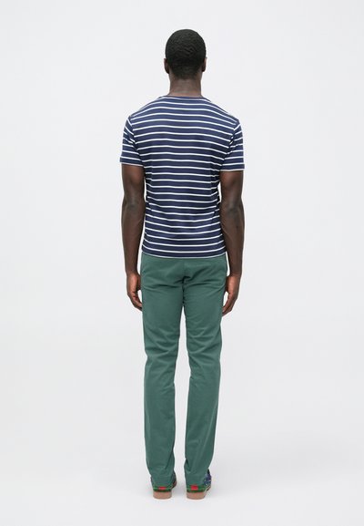 Tumši zila un balta svītraina īsroku krekls; zaļas bikses ar šauru piegriezumu; brūnas kurpes ar krāsainiem akcentiem. Skats no muguras.