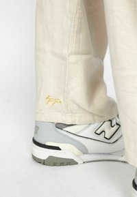 Beige vászonnadrág, melyen egy kis sárga hímzett logó található a szegélyen, szürke-fehér New Balance cipőkkel párosítva.