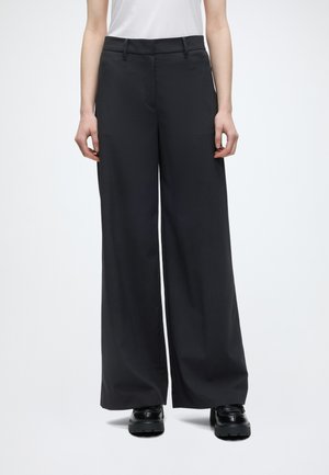 Person iført sort viddeben bukser, hvid indstukket top og sorte chunky plateau sko, der står mod ensfarvet baggrund.