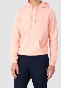 Felpa rosa chiaro con tasca frontale, cappuccio regolabile e logo ricamato sul petto, abbinata a pantaloni blu scuro.