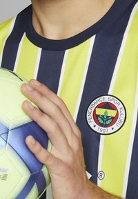 Žlutý a námořnický pruhovaný sportovní dres s emblémem Fenerbahçe. Osoba drží zeleno-modrý fotbalový míč. Textura vypadá prodyšně a lehce.