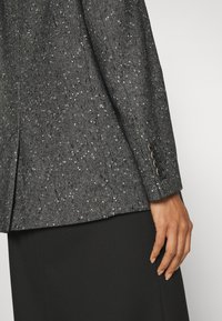 Gros plan sur le bras d'une personne portant un blazer gris foncé moucheté avec quatre boutons sur le poignet et une jupe noire, sur fond uni.