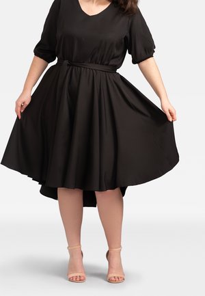 Frau trägt ein schwarzes knielanges Kleid mit Puffärmeln und beige-farbene Knöchelriemenabsätze, hält den Rocksaum seitlich ausgestreckt.