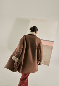 Cappotto oversize marrone con colletto e polsini in pelliccia sintetica bianca, indossato sopra pantaloni color ruggine, su uno sfondo minimalista.