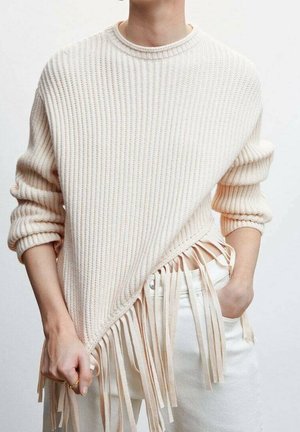 Personne portant un pull crème côtelé avec des manches retroussées et des détails à franges, associé à un pantalon blanc, tenant une frange du pull à la main.