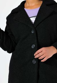 Manteau noir en mélange de laine texturé, doté d'un col cranté, de trois grands boutons et d'une coupe décontractée. L'intérieur laisse apparaître une chemise violette en dessous.