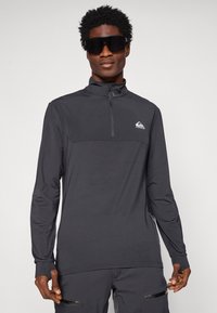 Mustade pika varrukaga pullover, millel on veerand tõmblukk, tekstureeritud kangas, kontrastne paneel rinna piirkonnas ja väike logo vasakul.