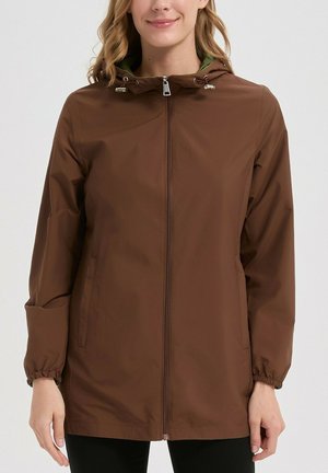 Femme portant une veste marron à capuche avec une fermeture éclair à l'avant et des poignets élastiques, debout devant un fond clair uni.