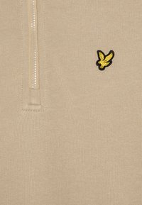 Μπεζ πουλόβερ με φερμουάρ ενός τετάρτου και ραβδωτό γιακά; διαθέτει ένα μικρό λογότυπο ενός μαύρου και κίτρινου πουλιού στο αριστερό στήθος. Λείος υφή.