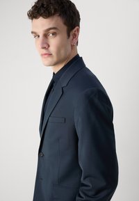 Giacca da abito blu navy con un design strutturato, chiusura a bottone singolo e rever a taglio. Sottile texture e una tasca a filetto sul petto.