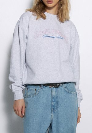 Szara bluza z oversize'owym krojem, z napisem "LOKALNI BOHATEROWIE" w różowym i niebieskim kolorze. Połączona z jasnoniebieskimi dżinsami i czarnym paskiem.