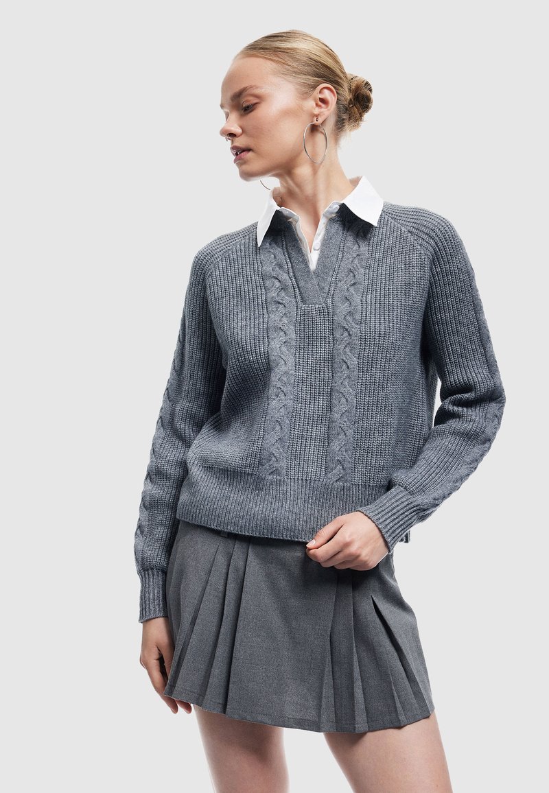 Szary sweter z dzianiny z dekoltem w serek, ozdobiony wzorami w warkocz, noszony na białej koszuli z kołnierzykiem. Połączony z szaro-plecioną spódnicą.