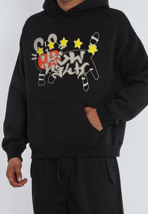 Hombre con sudadera negra con capucha con diseño abstracto de animal, cinco estrellas amarillas y texto estilizado en el frente, una mano en el bolsillo canguro.