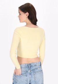 Top scurt cu mânecă lungă, ribbed galben, cu decolteu rotund, design ajustat și textură fină, purtat cu pantaloni scurți din denim cu talie înaltă.