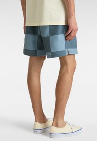 Blaue karierte Badeshorts mit einer leichten, glänzenden Oberfläche, kombiniert mit cremefarbenen Canvas-Sneakers mit weißen Schnürsenkeln und blauen Akzenten.
