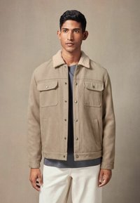 Jachetă bej din suede, cu un guler moale din shearling, două buzunare pe piept și nasturi negri, purtată peste o cămașă gri și o pereche de pantaloni albi.