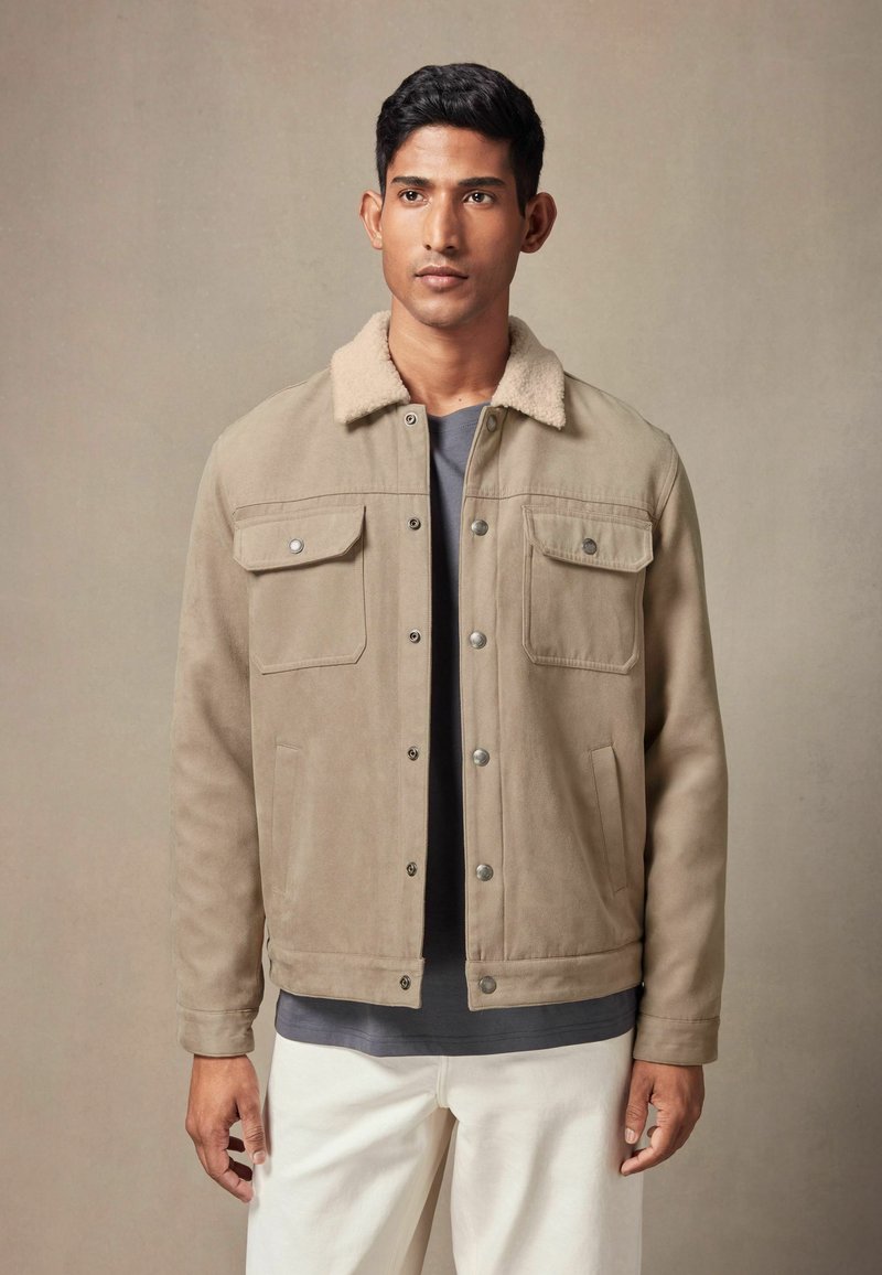 Beige Wildlederjacke mit einem weichen Schaffellkragen, zwei Brusttaschen und schwarzen Knöpfen, getragen über einem grauen Hemd und weißen Hosen.