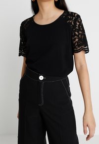 Svart t-shirt med spets på korta ärmar, rund halsringning, tillsammans med högmidjade svarta byxor med vit söm och knappstängning.