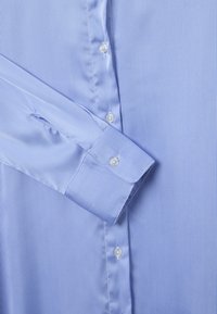 Camisa de botón a rayas azul claro con cuello estándar, puño simple y botones blancos, que presenta finas líneas verticales y una textura suave.